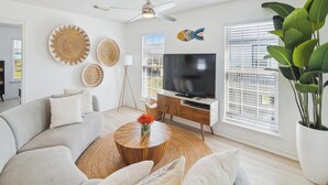 A modern living room with a sofa, coffee table, TV, decorative wall art, and a ceiling fan. - Seagull Sands by Ryson Vacation Rentals (Galveston)