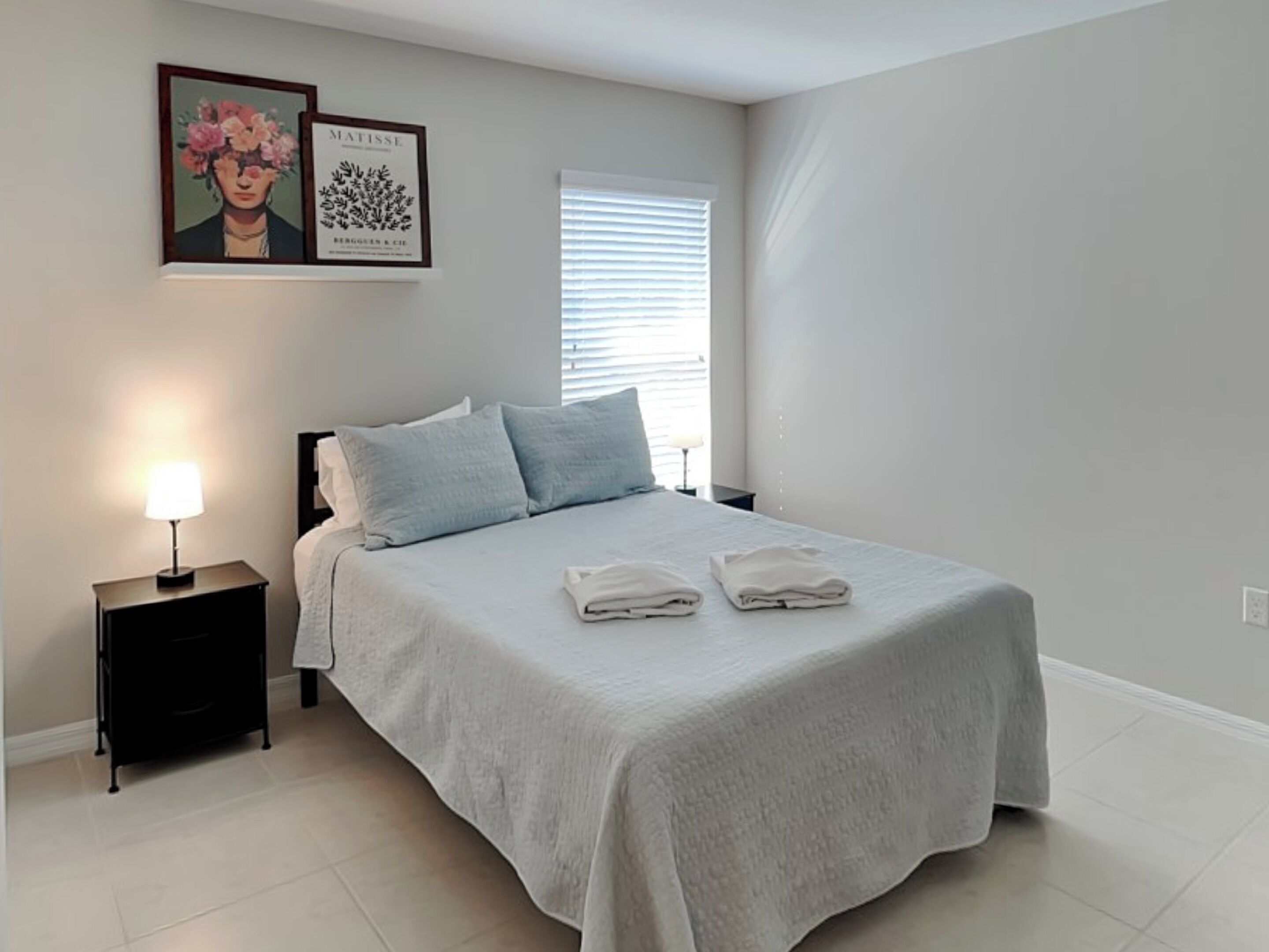 Una cama bien hecha con dos almohadas y dos toallas dobladas, una mesita de noche con una lámpara, y una obra de arte enmarcada en la pared.