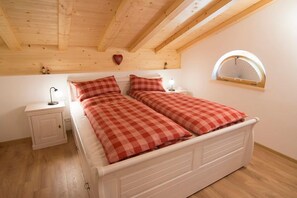 Ein Schlafzimmer mit karierter Bettdecke, einem Nachttisch mit Lampe, einem Fenster mit Ventilator und einer Holzbalkendecke.