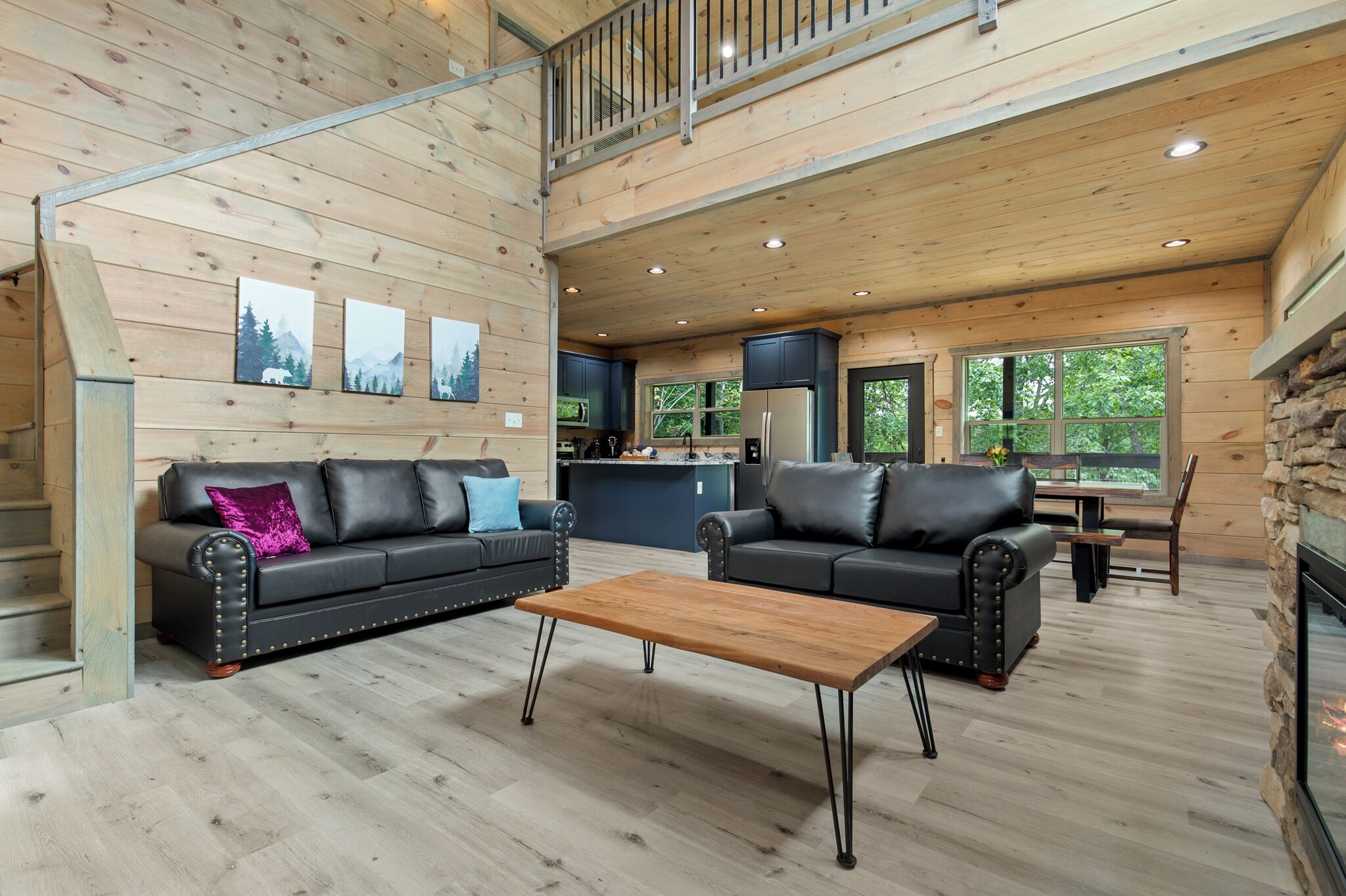 A spacious living area with a wooden floor, two black leather sofas with cushions, a wooden coffee table, and a stone fireplace.