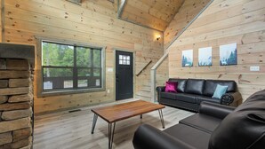 L’intérieur chaleureux d’un chalet avec une cheminée en pierre, une table à manger en bois, un canapé en cuir noir et une grande fenêtre donnant sur le paysage extérieur.