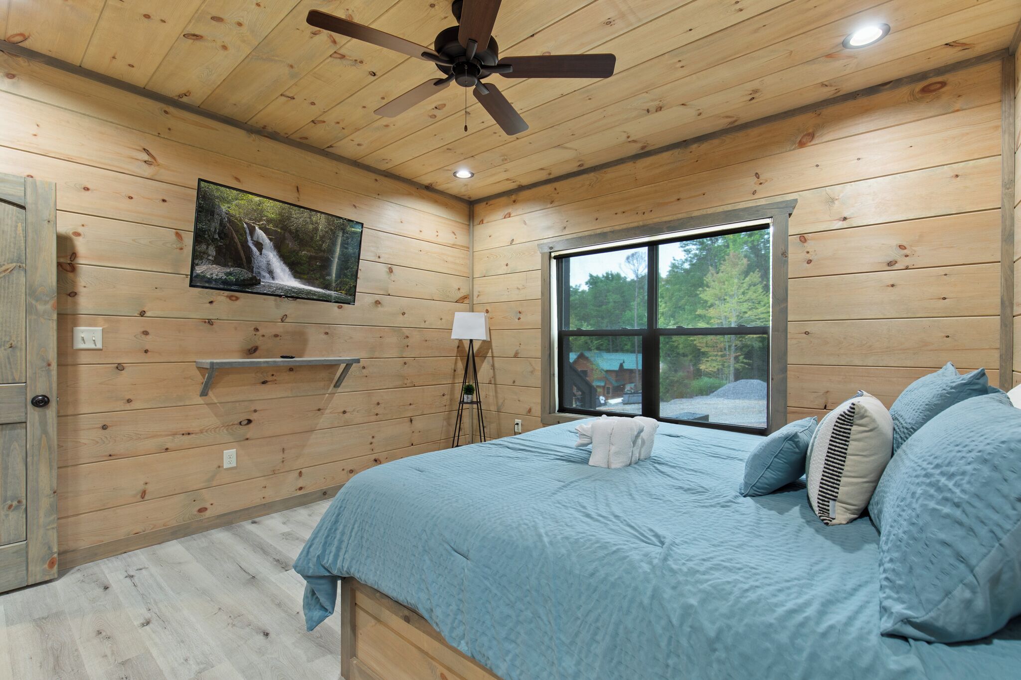 Ein Schlafzimmer einer Holzhütte mit Bett, Deckenventilator, Fernseher und Fenster mit Blick auf Bäume.