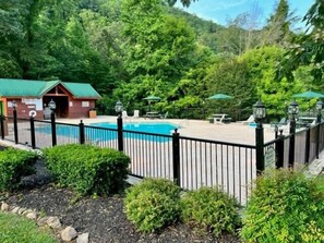 A pool area with a wooden cabin, surrounded by lush greenery and a black metal fence. - Splashin and Relaxin by Smoky Cove Resort (Sevierville)