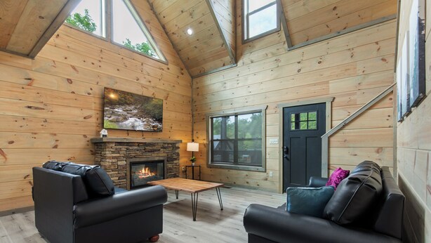 L’intérieur d’un chalet chaleureux, avec une cheminée en pierre, des murs en bois et deux canapés noirs.