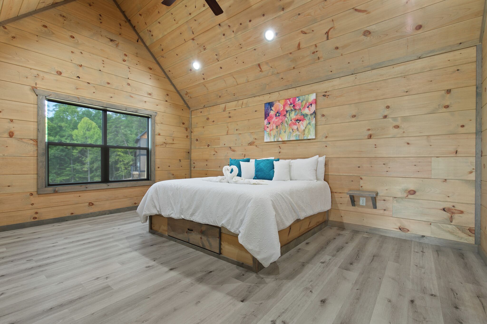 A wooden-paneled bedroom with a large window, a bed with white bedding and blue pillows, and a painting on the wall.