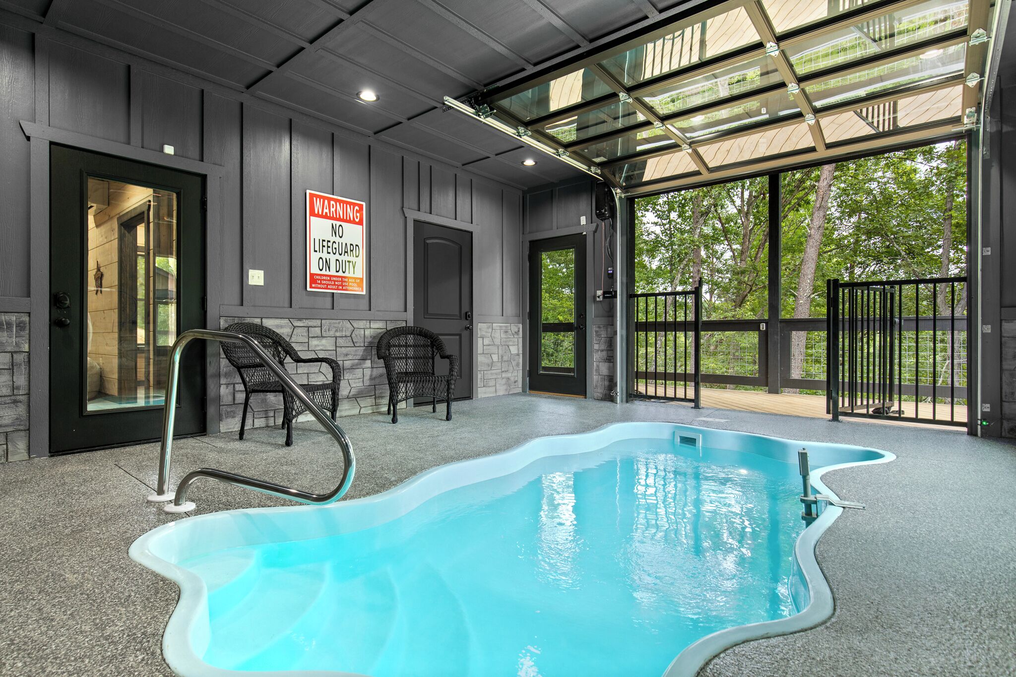Un área de piscina interior con un cartel de socorrista de servicio, dos sillas y un patio de cristal con vistas a árboles.