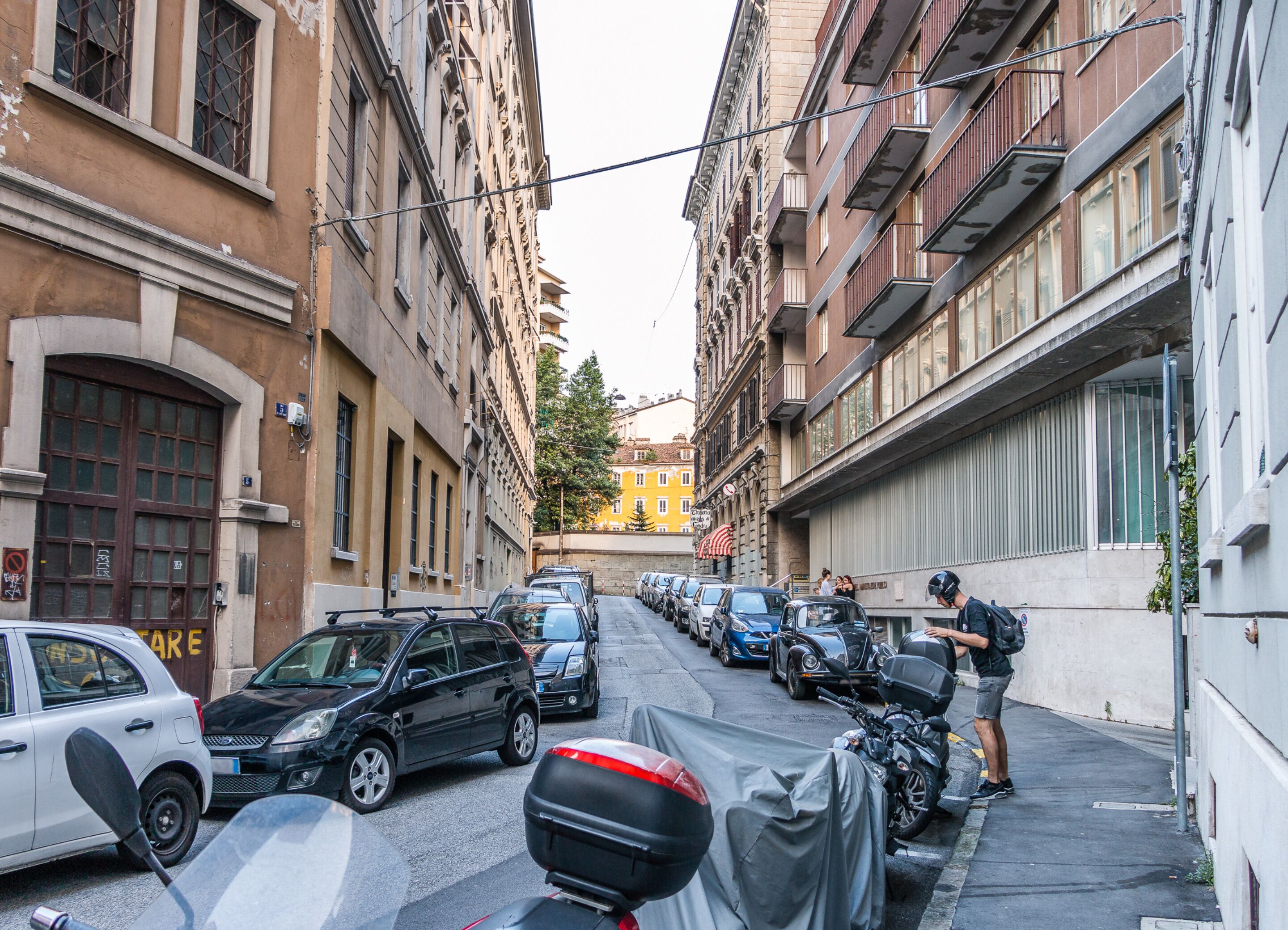 A narrow urban street lined with parked cars and motorcycles, featuring historic buildings with classic architecture.