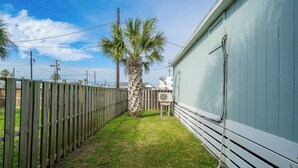 A fenced backyard with a palm tree, a utility box, and a building with a white exterior. - Tranquil Cove by Jackson Mountain Rentals (Port Aransas)