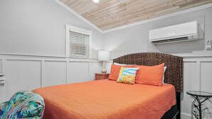 A bedroom with a wooden ceiling, white walls, an air conditioner, a bed with an orange bedspread, a lamp, and a small table. - Tranquil Cove by Jackson Mountain Rentals (Port Aransas)