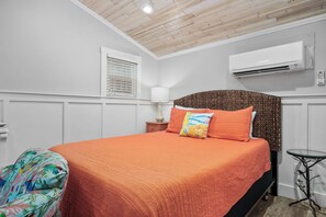 A bedroom with a wooden ceiling, white walls, an air conditioner, a bed with an orange bedspread, a lamp, and a small table. - Tranquil Cove by Jackson Mountain Rentals (Port Aransas)