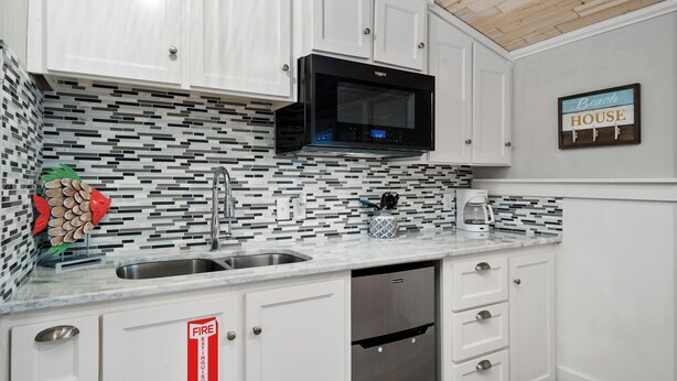 A kitchen with white cabinets, a black microwave, a stainless steel sink, and a tiled backsplash. - Tranquil Cove by Jackson Mountain Rentals (Port Aransas)