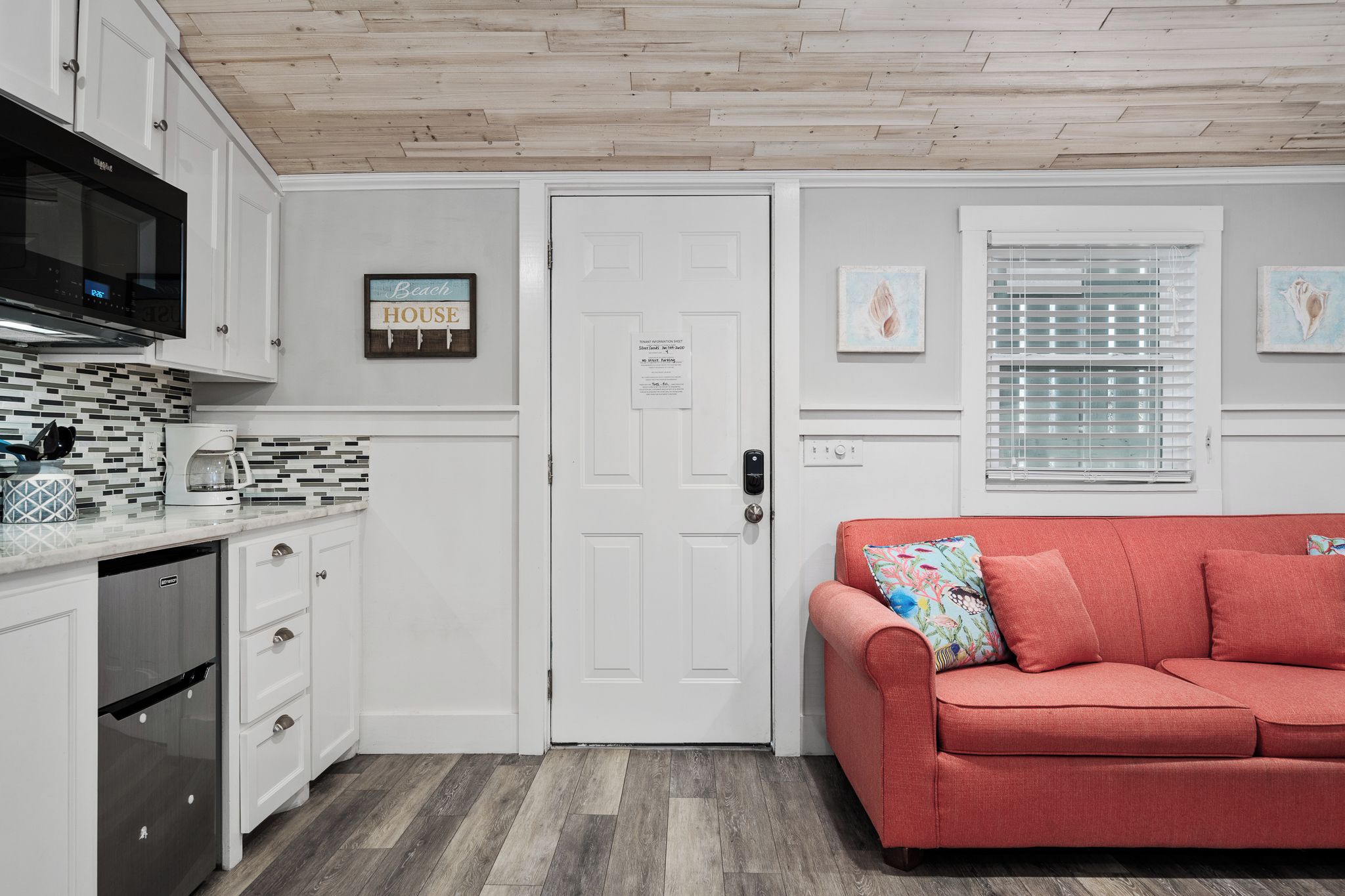 A compact kitchen with white cabinetry, a microwave, and a coffee maker. There's a door with a notice, a window with blinds, and a red sofa with a colorful pillow.