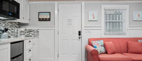 A compact kitchen with white cabinetry, a microwave, and a coffee maker. There's a door with a notice, a window with blinds, and a red sofa with a colorful pillow.