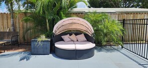 Uma área de estar circular ao ar livre, com toldo, cercada por um muro de madeira e vegetação exuberante.
