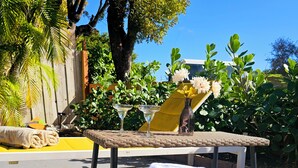 Un área de estar al aire libre iluminada por el sol, con una mesa de mimbre, dos sillones y un jarrón con flores.