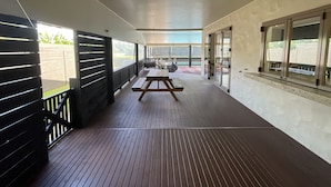 Un espacio al aire libre techado con terraza de madera, una mesa de picnic y vistas al jardín a través de amplios ventanales.