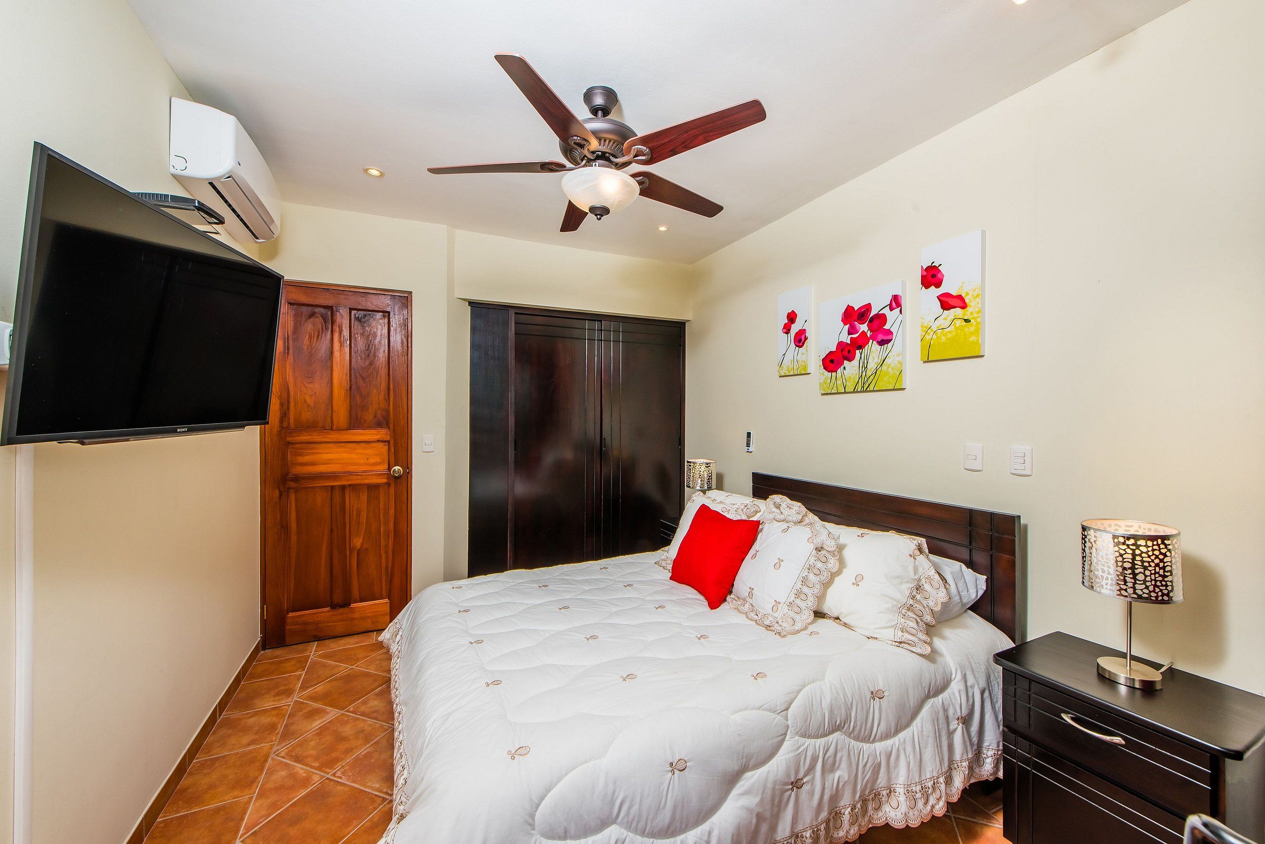 A bedroom with a bed, a ceiling fan, a TV, a wardrobe, and artwork on the wall.