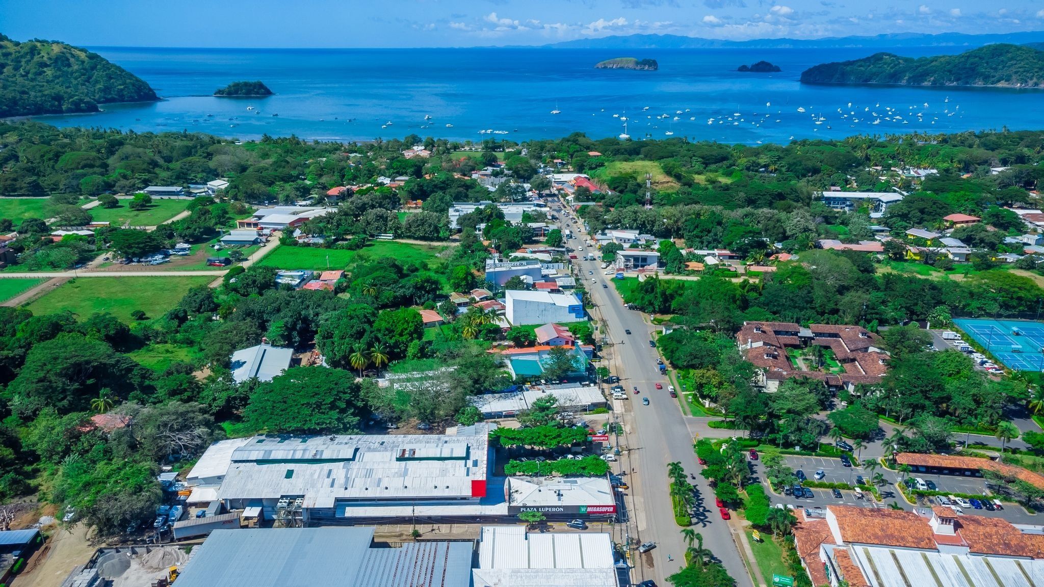 An aerial view of a coastal town with a main road, buildings, and a marina with boats.