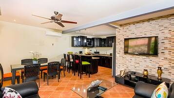 A modern living room with a dining area, kitchen, and a large TV mounted on a brick wall.
