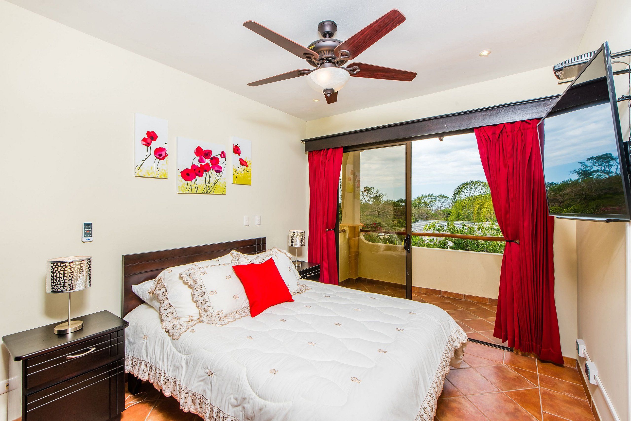 A bedroom with a bed, a nightstand, a ceiling fan, a window with red curtains, and a balcony.