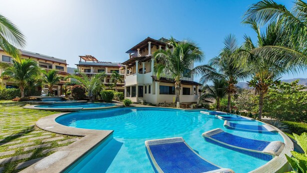 A large outdoor swimming pool with multiple lounge areas, surrounded by palm trees and a multi-story building. - Coco Sunset Hills 52 (Coco)