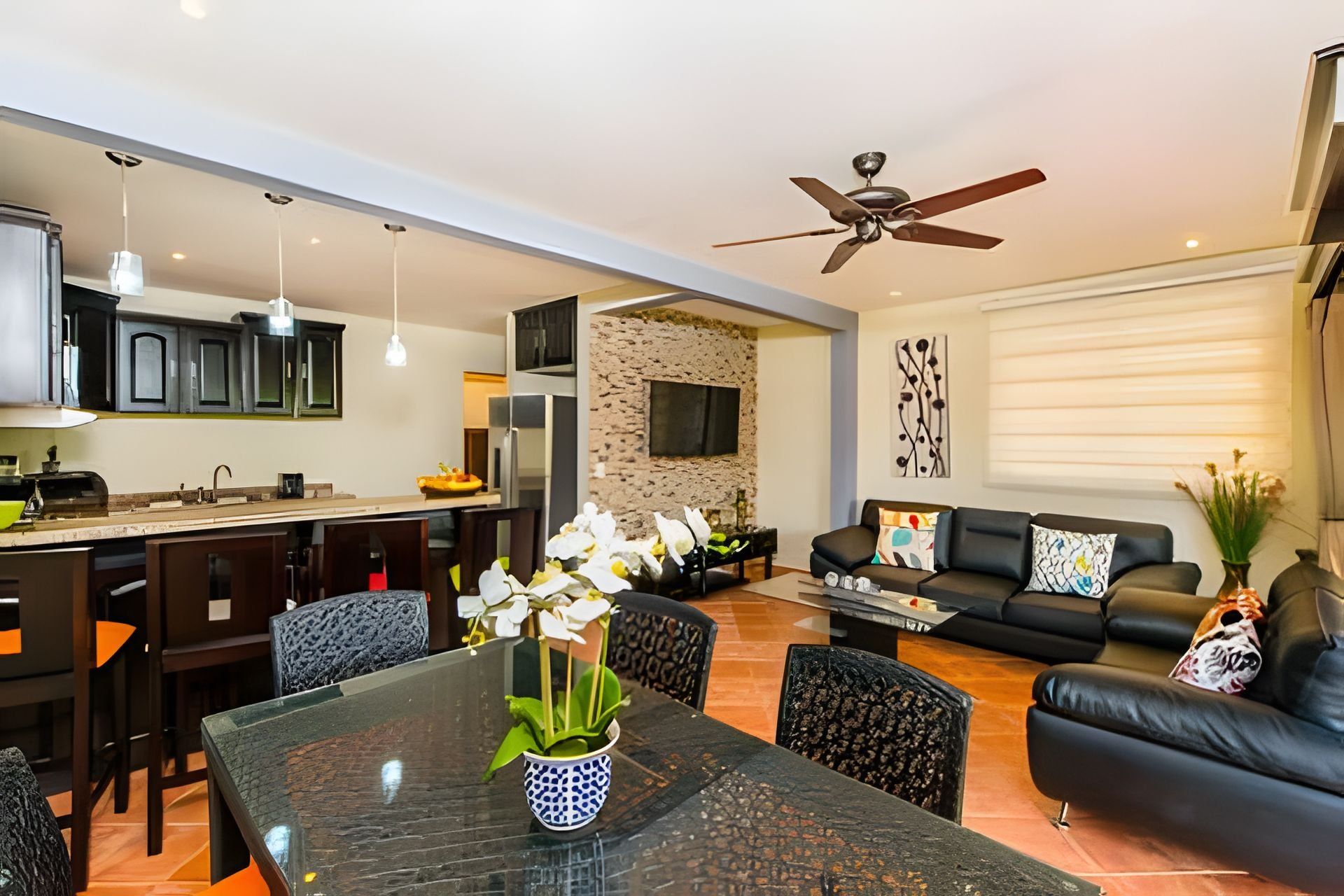 A modern kitchen with a dining area, a living room with a sofa and a TV, and a ceiling fan.