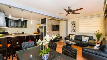 A modern kitchen with a dining area, a living room with a sofa and a TV, and a ceiling fan.