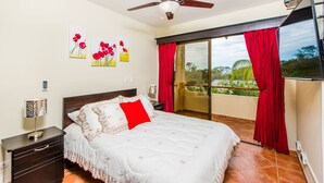 Un dormitorio con cama, mesita de noche, ventilador de techo, ventana con cortinas rojas y balcón.