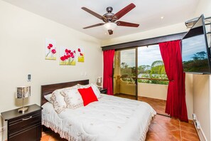 Un dormitorio con cama, mesita de noche, ventilador de techo, ventana con cortinas rojas y balcón.