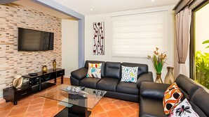 A modern living room with a stone wall, a black leather sofa, a glass coffee table, and a flat-screen TV mounted on the wall. - Coco Sunset Hills 52 (Coco)