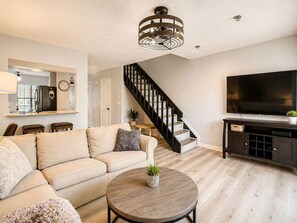 Una sala de estar con sofá, una mesa de centro, televisor, escalera y una zona de cocina.