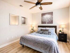Un dormitorio con cama, dos mesitas de noche con lámparas, ventilador de techo y una obra de arte en la pared.