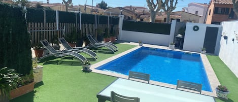 A rectangular swimming pool with a tiled edge, surrounded by artificial green turf. There are lounge chairs and a table with chairs in the foreground.