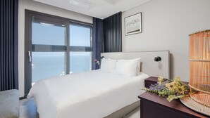 A hotel room with a large bed, a nightstand with a vase of flowers, a view of the sea through a large window, and a framed picture on the wall. - NewCZ Osiria Residence (Busan)