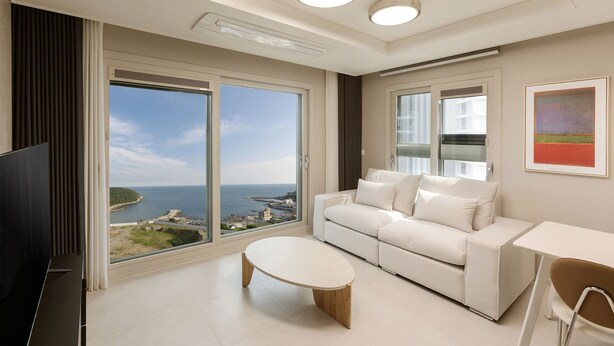 A modern living room with a white sofa, a small round table, and a TV. It features large windows with a view of the sea and a building. - NewCZ Osiria Residence (Busan)