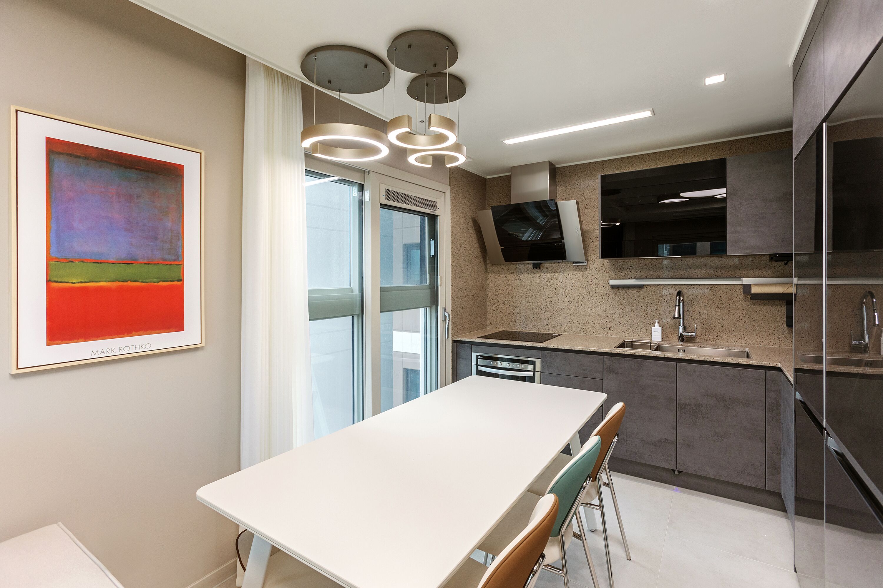 A modern kitchen with a white table, bar stools, a large painting, and a TV mounted on the wall.
