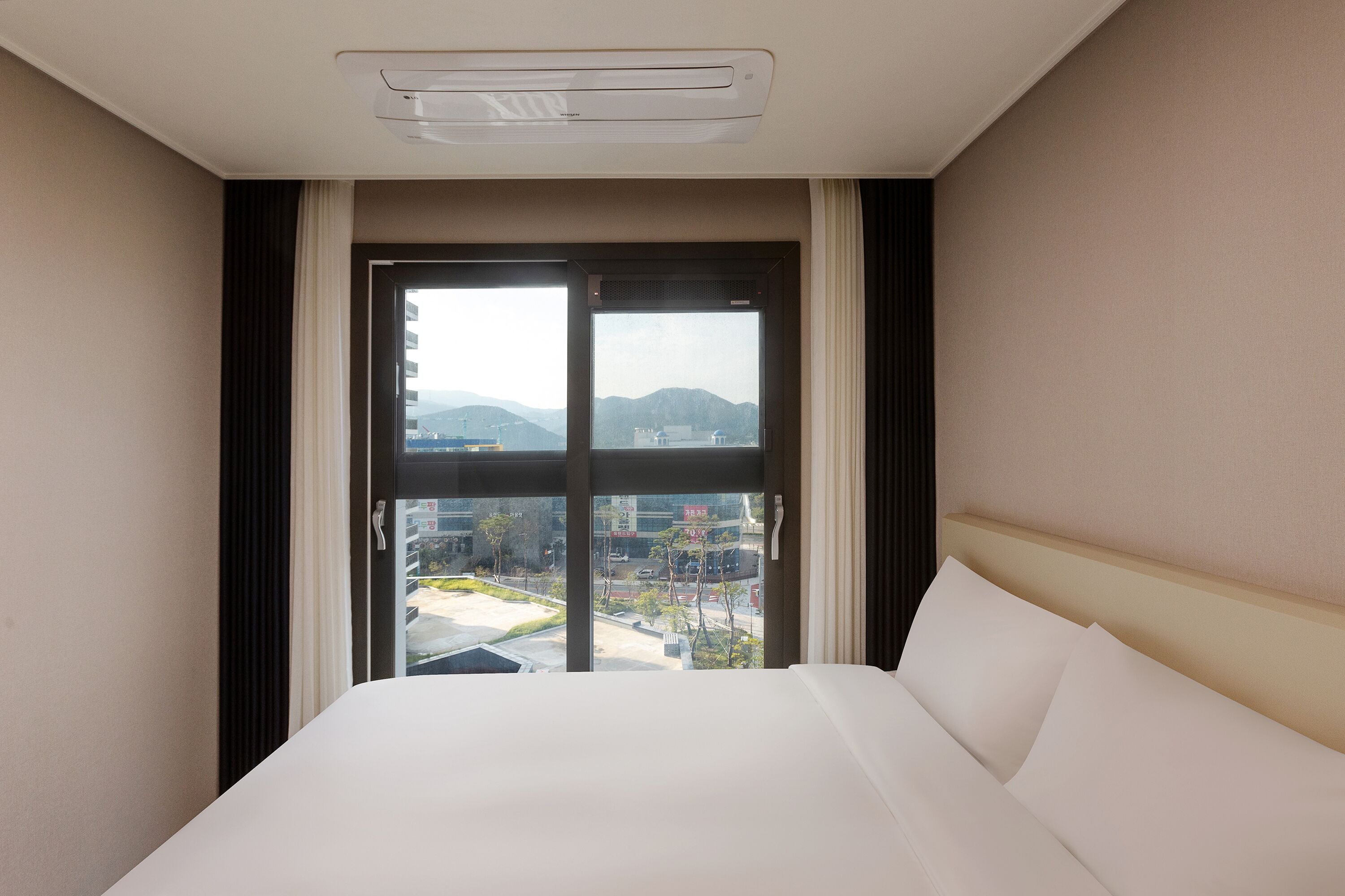 Et hotelværelse med en stor seng, et vindue der viser bygninger og bjerge, og en aircondition i loftet.