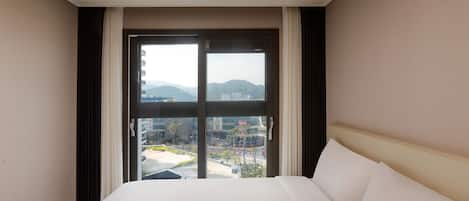 A hotel room with a large bed, a window with a view of buildings and mountains, and a ceiling air conditioner.