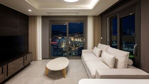 A modern living room with a flat-screen TV, a sofa, a coffee table, and a city view through large windows. - NewCZ Osiria Residence (Busan)