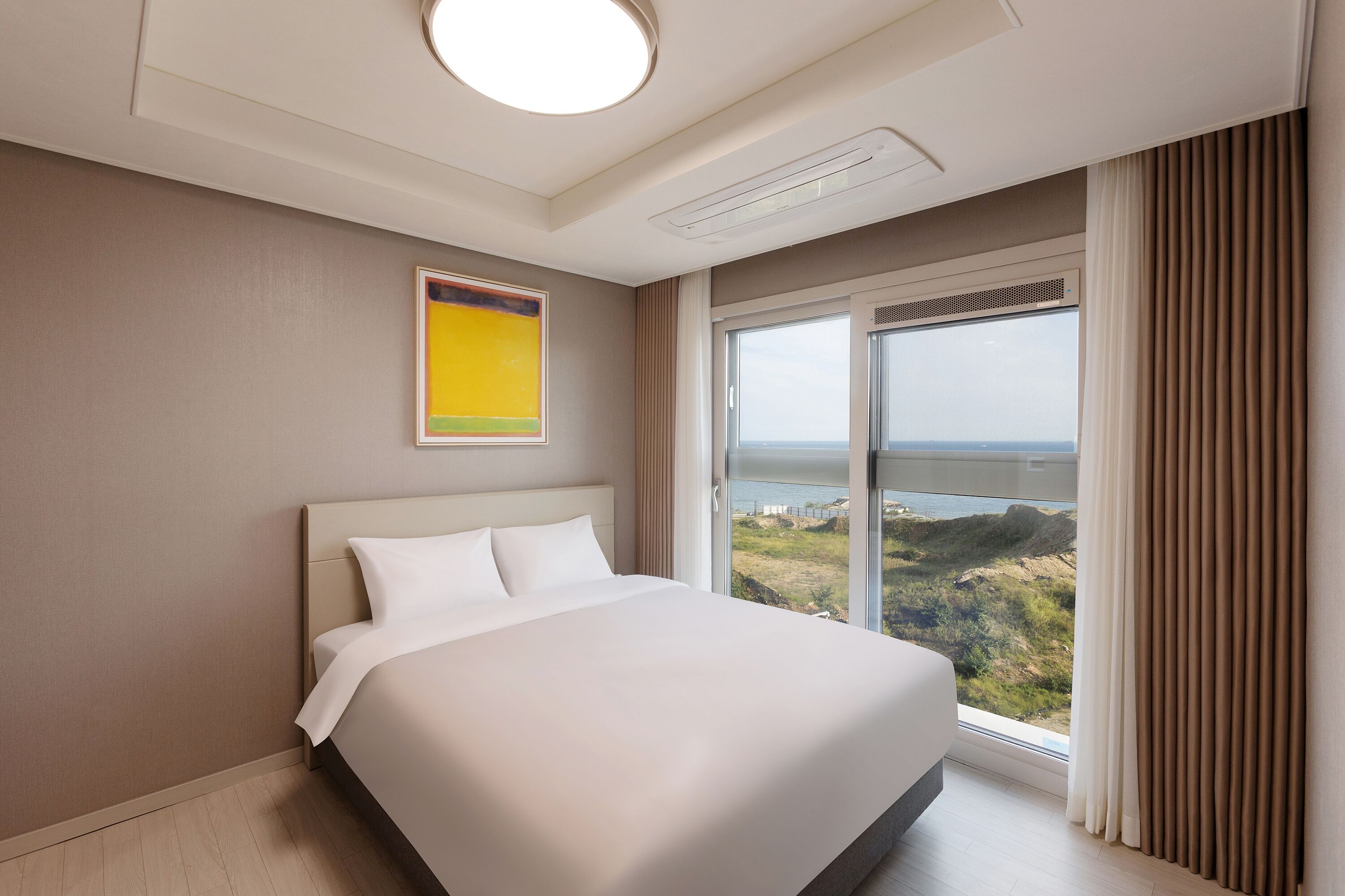 A modern hotel room with a large bed, a view of the landscape through a window, and a ceiling light.