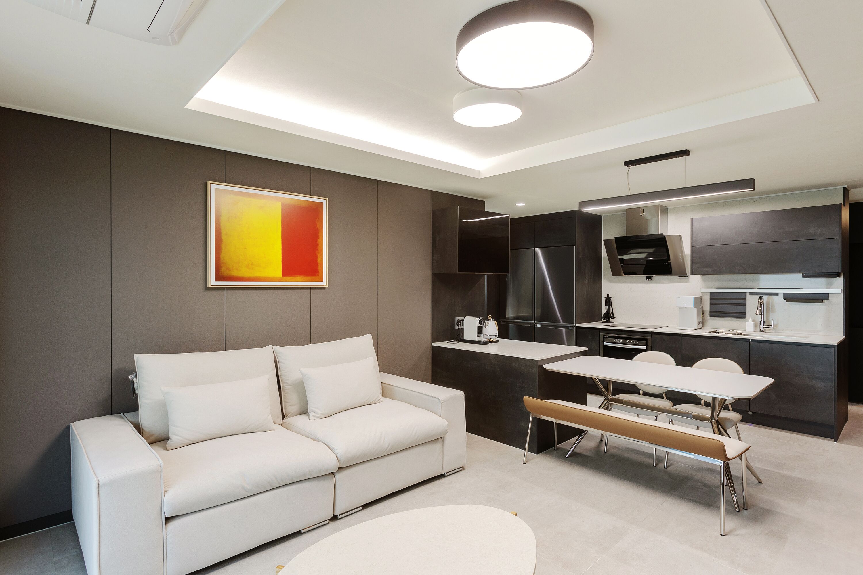 A modern living room with a sofa, a dining table, and a kitchen area with dark cabinetry and a stainless steel refrigerator.