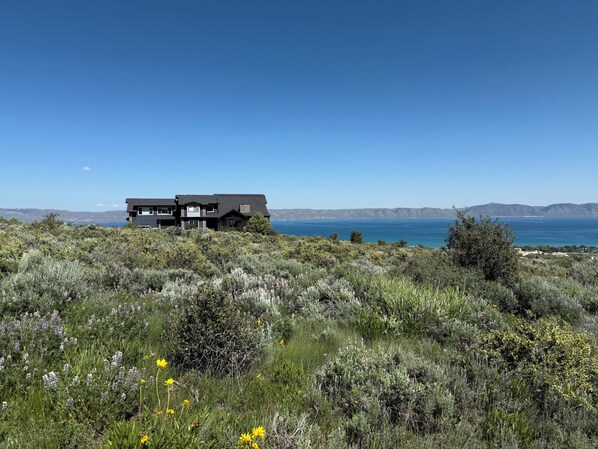 A modern two-story house with a dark roof, situated on a grassy hillside overlooking a coastal landscape with mountains in the distance. - Spectacular views of Bear Lake in a spacious private retreat on nearly 6 acres! (Fish Haven)