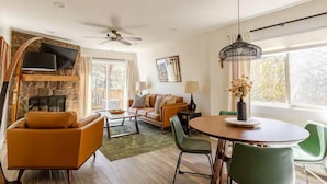 A modern living room with a stone fireplace, a ceiling fan, a television, and a dining area with a round table and chairs. - Quiet 2BR cottage, King Bed, Fire, <1 mi to Sugar (Avery County)