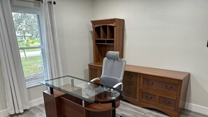 A modern office space with a glass desk, a grey office chair, and a wooden cabinet with drawers. - Beautiful family house in Venice, Fl. It is only a mile away from Shamrock Park  (Venice)