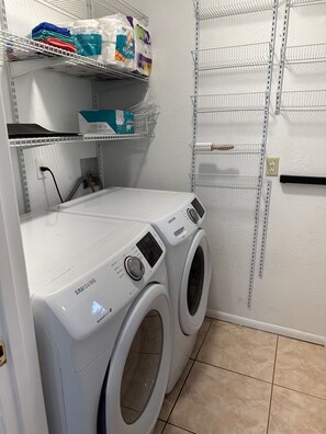 A laundry room with a Samsung washing machine and dryer, shelves with laundry supplies, and a tiled floor. - Beautiful family house in Venice, Fl. It is only a mile away from Shamrock Park  (Venice)