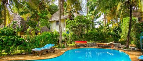 A swimming pool surrounded by lush greenery and a thatched-roof house.
