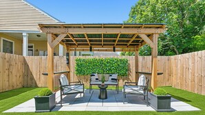 A covered patio area with a wooden pergola, a seating arrangement, and a green hedge.