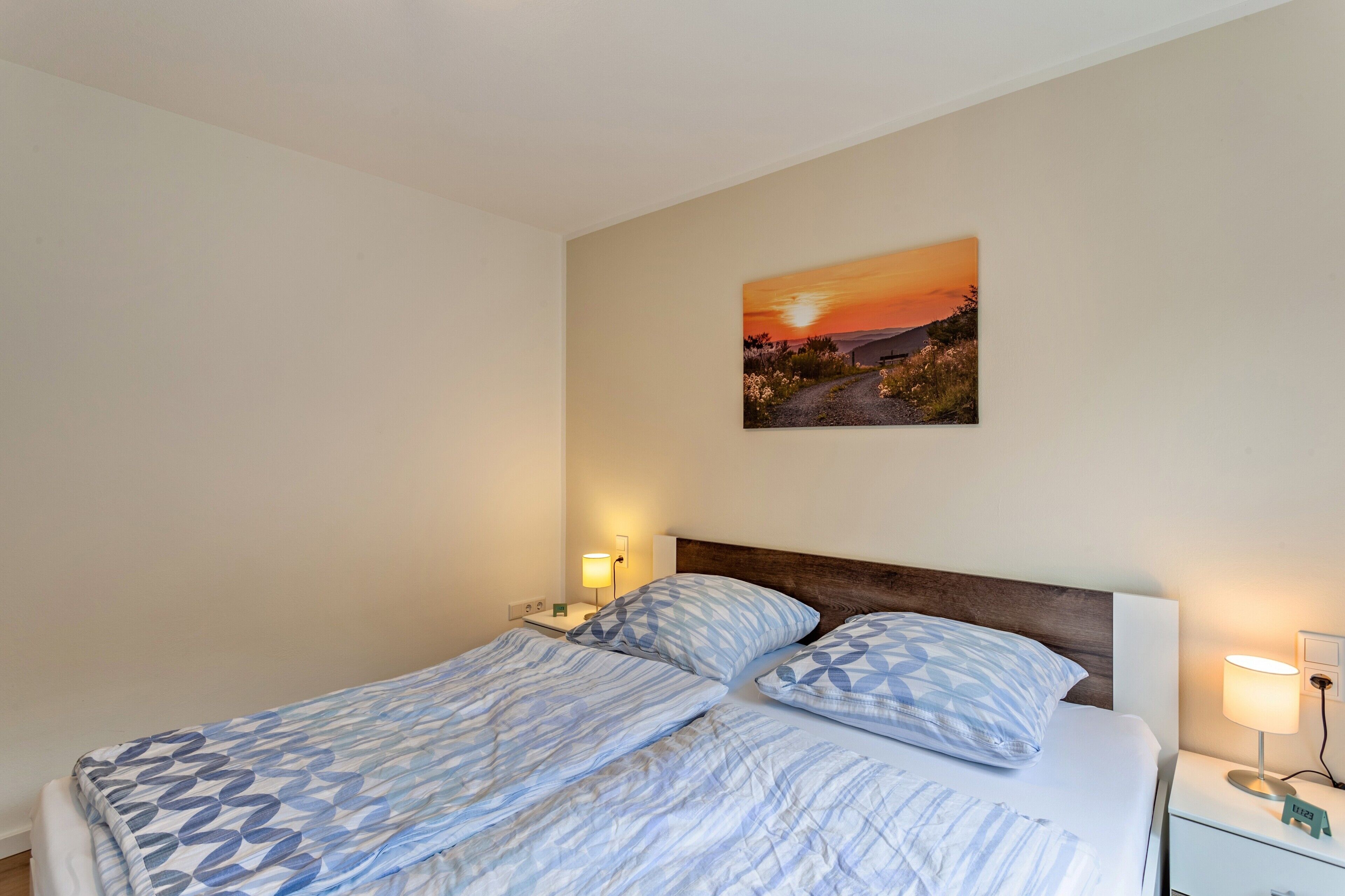 Una cama bien hecha con ropa de cama estampada, un cuadro colgado en la pared que representa un atardecer y lámparas de noche.