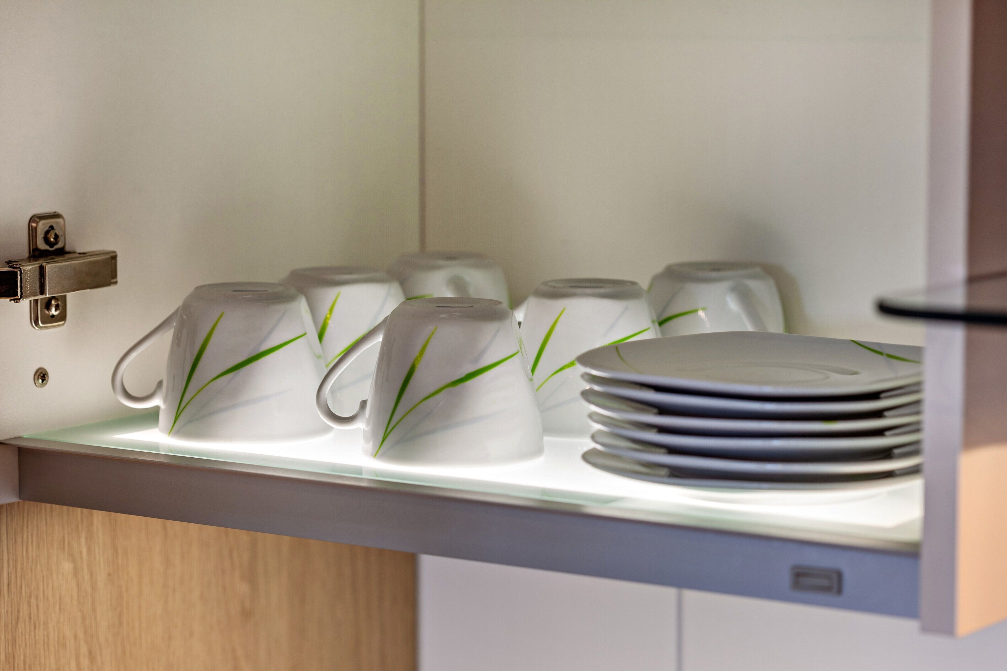 A kitchen area with a white countertop, a stack of white plates, and a row of white ceramic mugs with green accents.