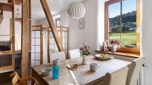 Een eethoek met een houten tafel, stoelen, een vaas met bloemen en uitzicht op een groen landschap door een raam.